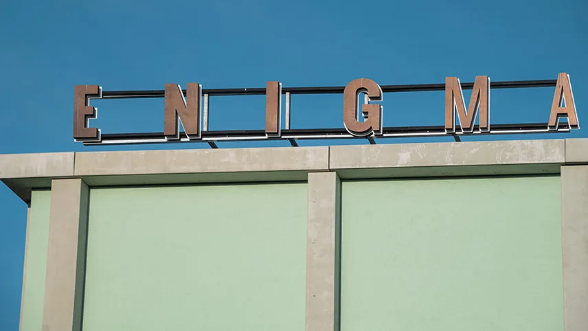 Byggnaden sedd utifrån med ljusgrön fasad och namnet ENIGMA i blockbokstäver på taket, mot klarblå himmel.