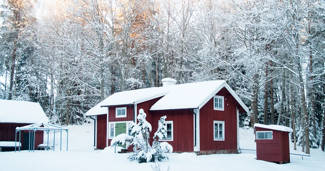 En rød skihytte i Sverige omgivet af sneklædte træer og et vinterlandskab, med et tykt lag sne på taget og jorden, hvilket skaber en idyllisk og fredfyldt vinterstemning.