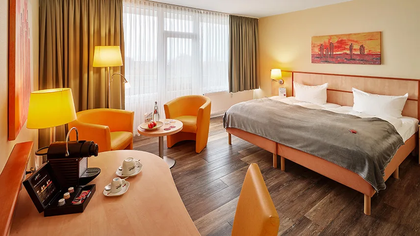 Bild av ett av rummen på strandhotellet. Man ser en dubbelsäng bäddad med vita kuddar och ett grått överkast, ett stort fönster med vita gardiner och en sittgrupp. I förgrunden syns ett bord med två framdukade kaffekoppar och en liten kaffemaskin.