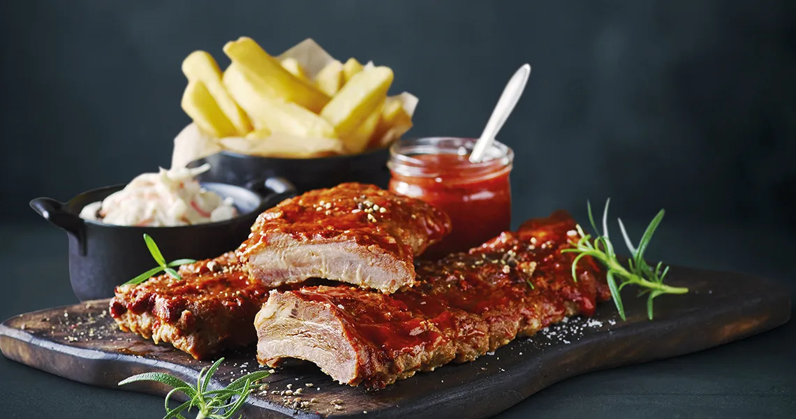 Revbensspjäll med barbecuesås ligger på en skärbräda framför en skål med pommes frites, en kopp med coleslaw och en burk med mer sås i en fotostudio.