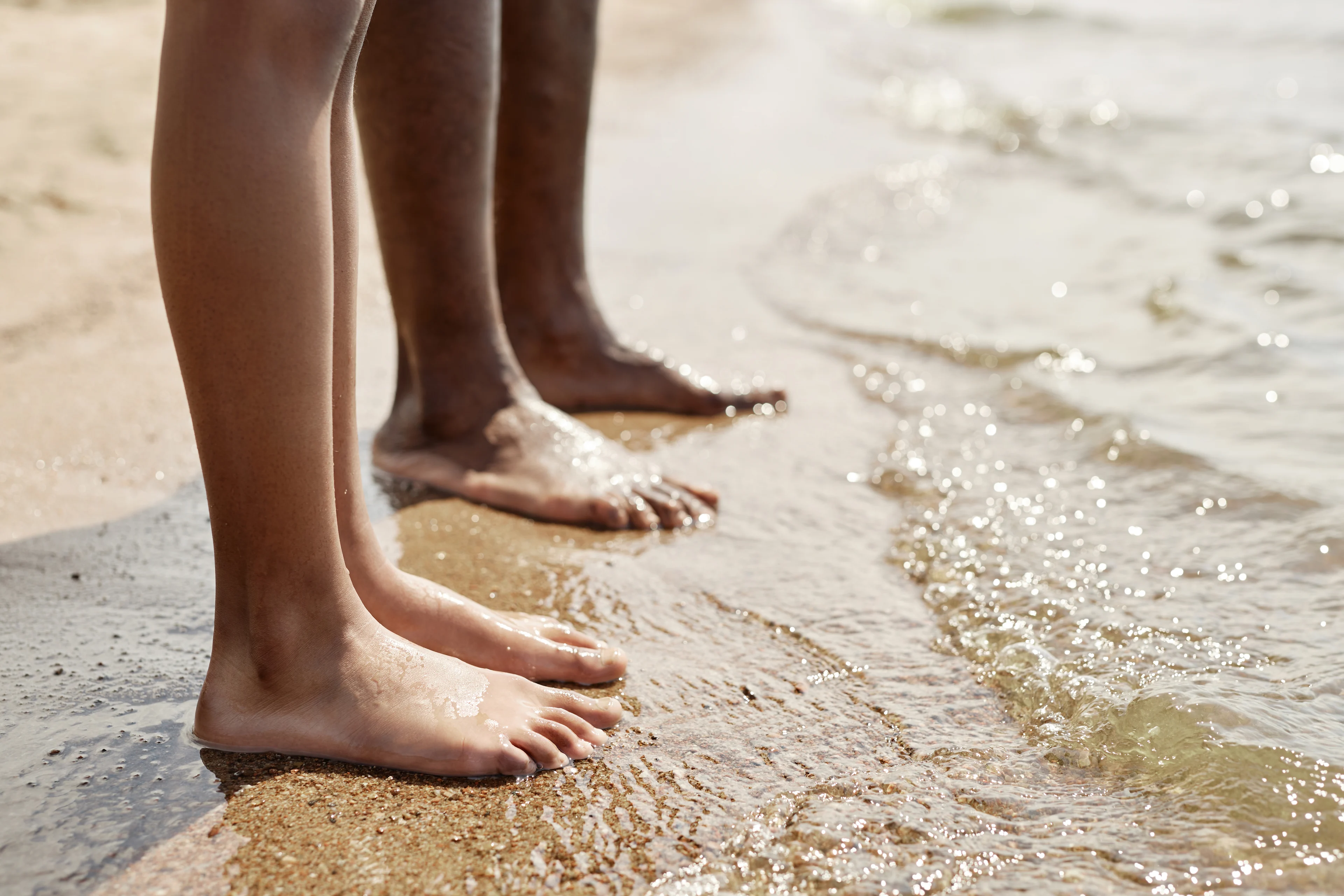 Barn och vuxen som står barfota i vattenbrynet på strand.