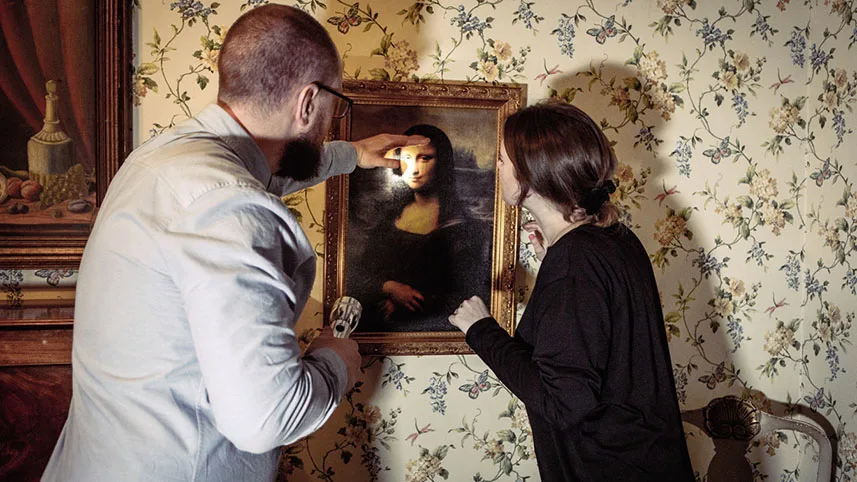 Två besökare står vid en vägg med blommig tapet och tavlan Mona Lisa. En av besökarna pekar mot tavlan och lyser med en ficklampa medan den andra tittar på.
