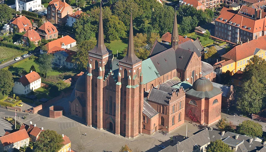 Flygfoto av Roskilde domkyrka. Man ser kyrkan snett uppifrån mot en bakgrund av gröna gräsmattor, grönskande träd och byggnader i tegel.