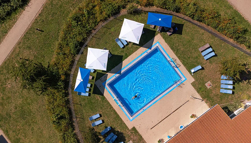 Flygbild av hotellets poolområde utomhus. I mitten ser man den stora utomhuspoolen och runtomkring syns parasoller och sittgrupper.