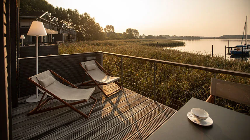 Utsikt från ett av semesterhusens veranda. Man ser två vilstolar och en lampa samt en kaffekopp på ett bord. I bakgrunden syns solen gå ner över vattnet.