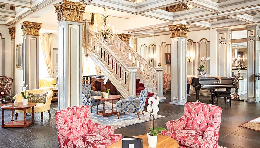 Hotellets vackra lobby med mörka trägolv och möbler i elegant stil. Mitt i rummet syns en bred trappa med pelare på sidorna och en kristallkrona lyser upp rummet.