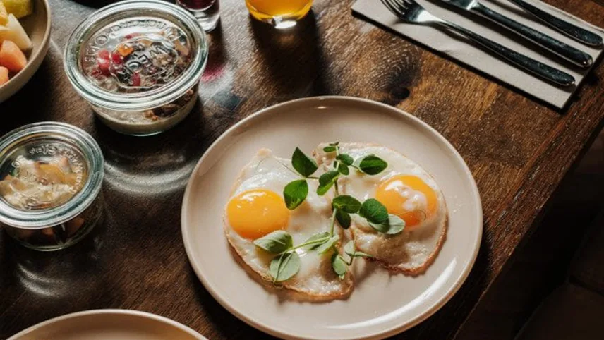 Bild från hotellets frukost. Här syns en beige tallrik med två stekta ägg med gulan upp och en grön kvist som pynt. Runt tallriken syns små glasskålar med frukt och yoghurt.