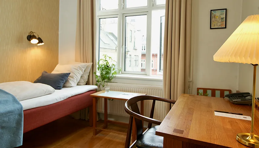 Bild från ett av hotellets 50-talsrum. Man ser en dagbädd med vackra textilier, ett skrivbord med stol och lampa samt ett fönster ut mot en innergård.