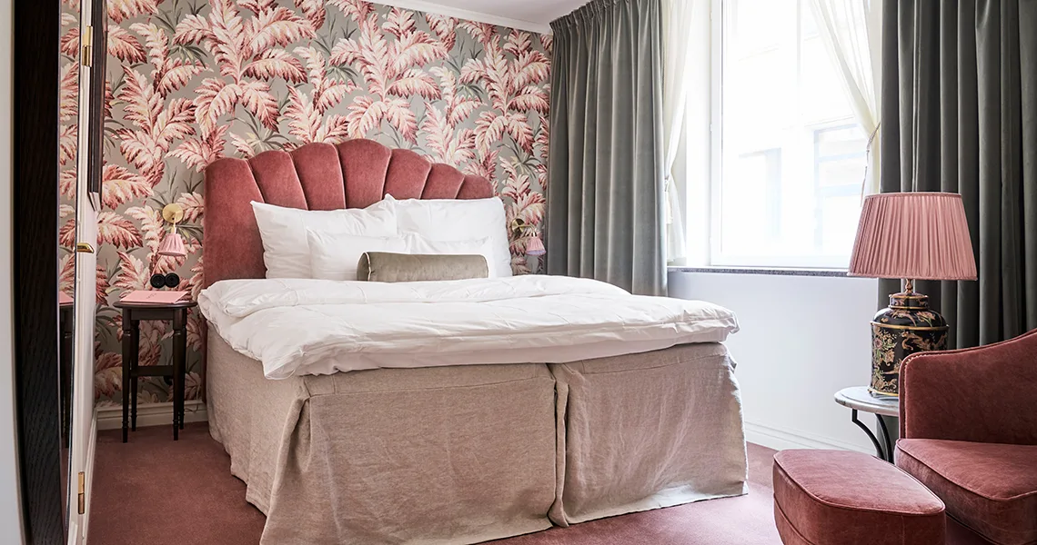 En seng med et hovedgærde i pink fløjl står i et rum med blomstret tapet, grønne gardiner og en pink lænestol.