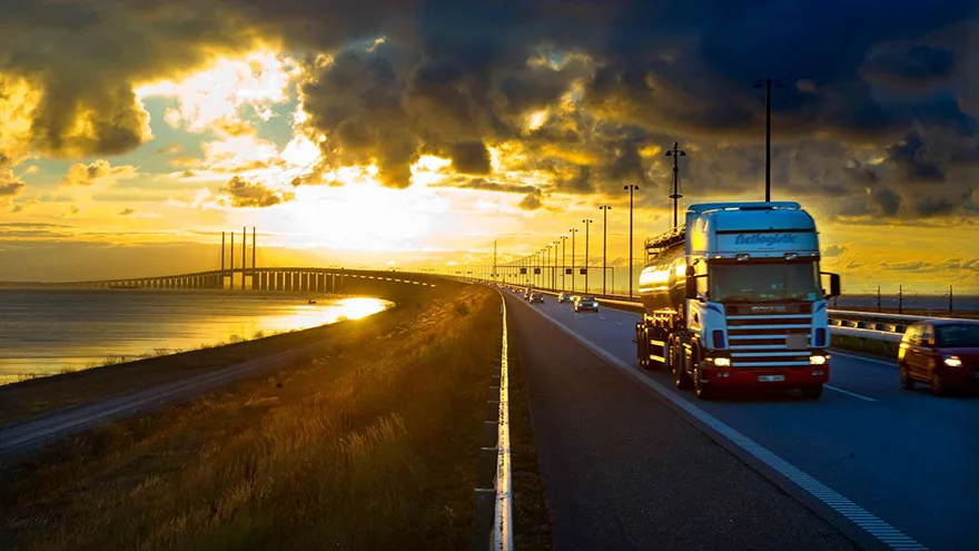 En tung lastbil på bron vid solnedgång. I bakgrunden syns högbron i solnedgång och en vacker himmel med stora moln.
