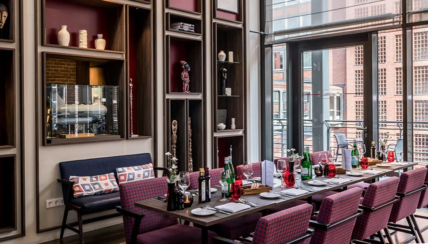 Hotellets restaurang cantinetta ristorande & bar med glasvägg ut mot en balkong, ett långbord dukat med tallrikar, färgglada glas och flaskor. Väggarna är klädda i trähyllor med prydnadssaker.