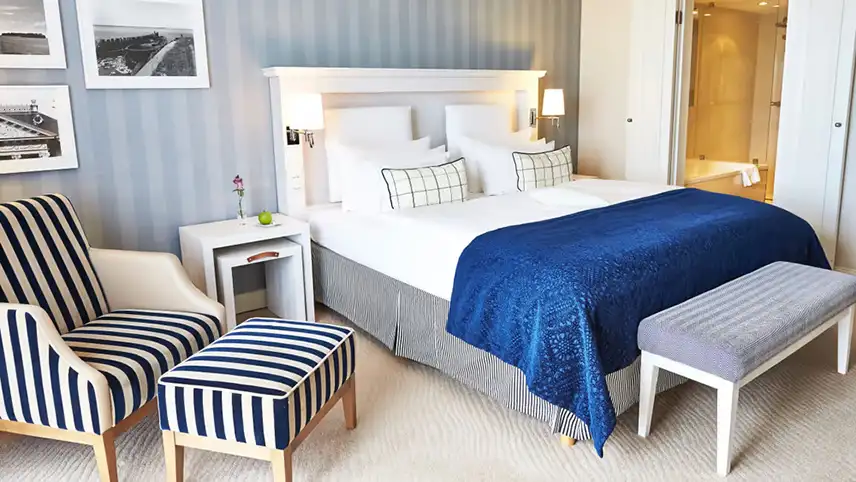 Ett dubbelrum med ljusblå väggar, stor dubbelsäng bäddad med vita och blå sängkläder och en charmig fåtölj med matchande fotpall i blå- och vitrandigt typ.