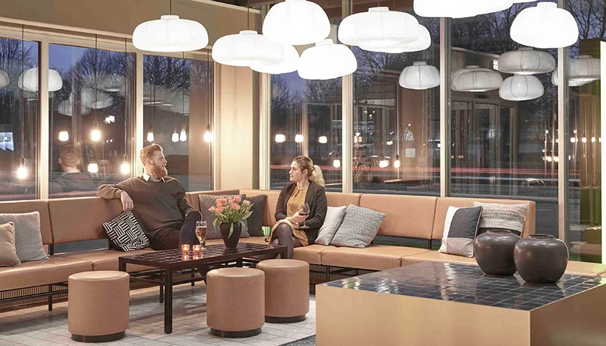 Loungeområde med stora sittgrupper och lampor i taket. Två personer sitter i en soffa med varsitt glas och pratar.