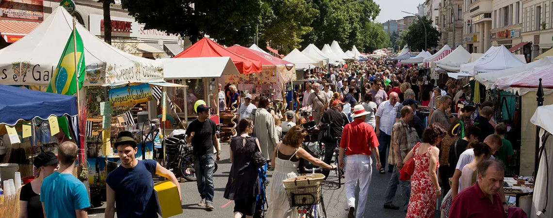 En livlig marknadsgata i Berlin fylld med människor och stånd som säljer olika varor, med färgglada tält och träd som skuggar området på en solig dag.