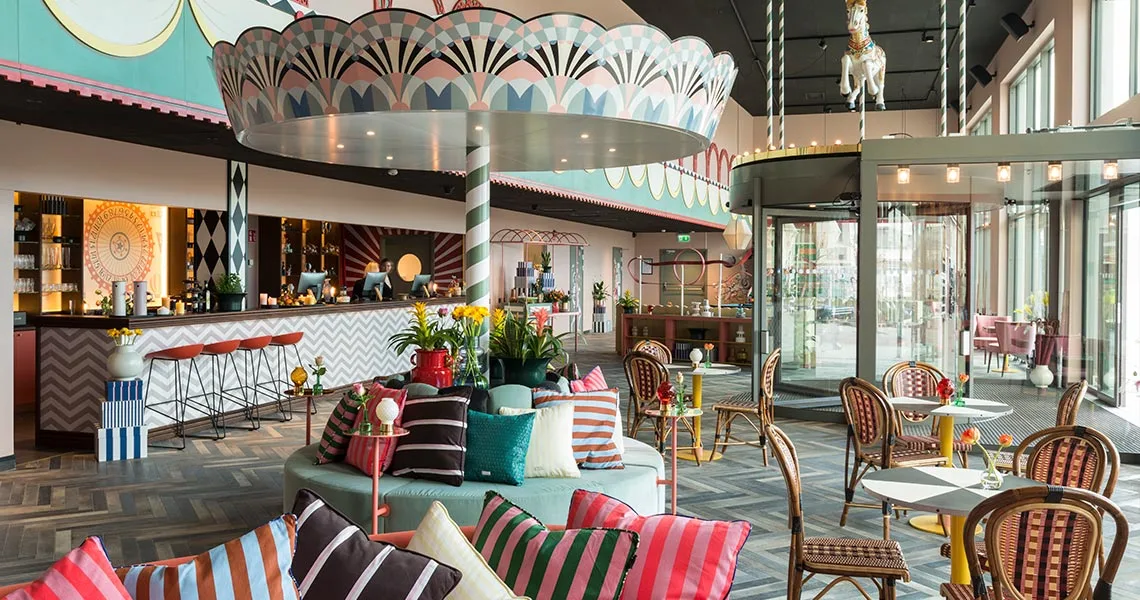 En farverig café med puder, stole og blomster på bordene ved siden af en oplyst bar og en karruselhest.