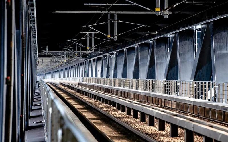 Øresundsbrons järnväg utan tåg, med dubbla stålspår och grå metallkonstruktioner som sträcker sig genom bron. Bilden visar en tom järnvägskorridor med bruna slipers, silverfärgade räls och svarta elledningar under ett mörkt tak, vilket framhäver den moderna infrastrukturen mellan Danmark och Sverige.