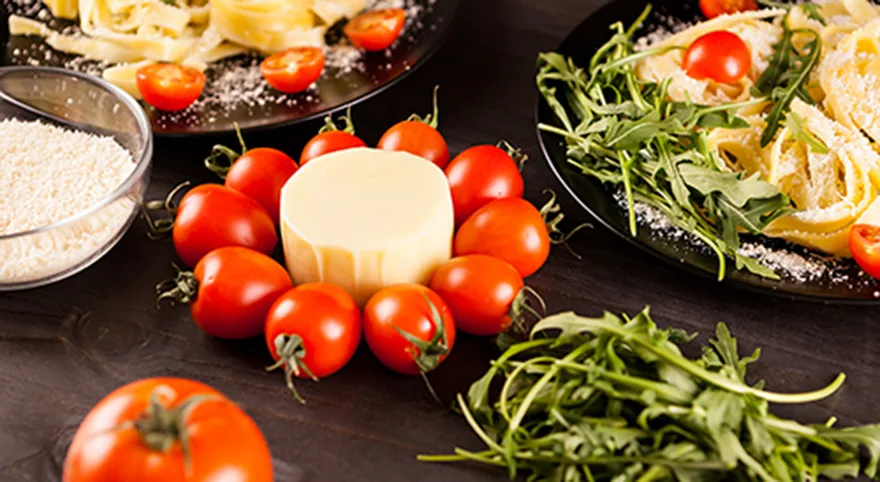 Närbild av färska röda tomater, en rund ost, ruccola och tallrikar med pasta.