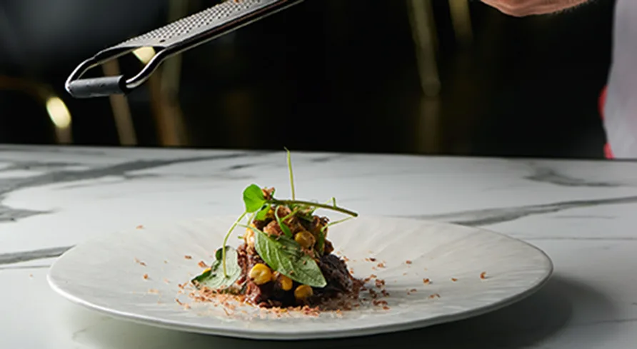 En tallrik står på en bordsyta i marmor och överst i bilden ser man en del av ett rivjärn som river tryffel över en liten rätt.