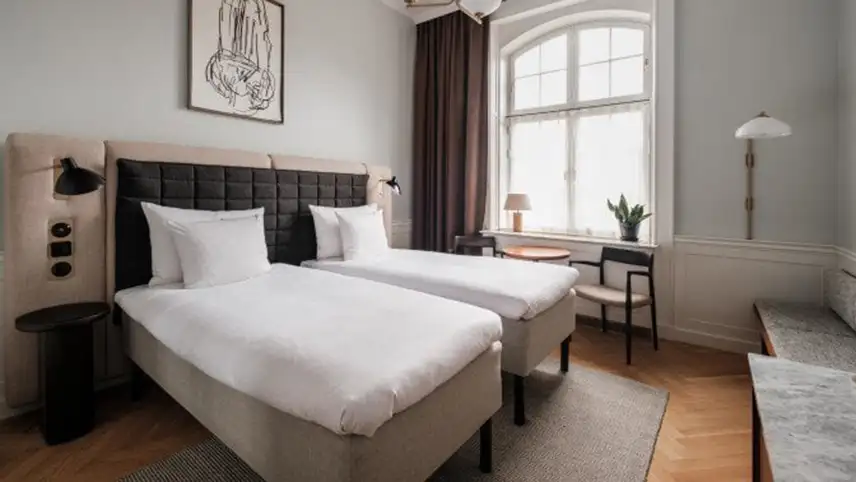 Ett av rummen med bred dubbelsäng med sänggavel i beige och svart, vita sängkläder samt fluffiga täcken.