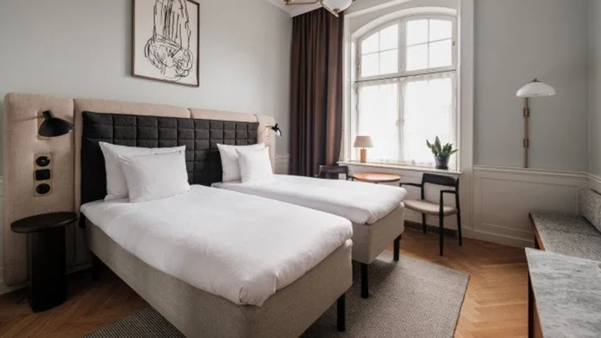 Ett av rummen med bred dubbelsäng med sänggavel i beige och svart, vita sängkläder samt fluffiga täcken.