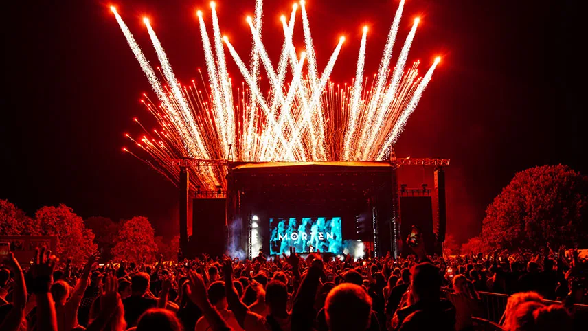 Konsert under förra årets festival. Det är kväll, scenen är upplyst av blått ljus och fyrverkerier på himlen. I förgrunden dansar publiken med armarna i luften.,