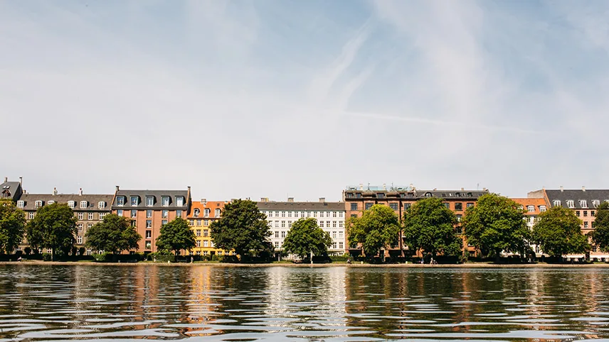 Lummiga lövträd längs en av sjöarna i centrala Köpenhamn. I förgrunden syns spegelblankt vatten och i bakgrunden en ljusblå sommarhimmel med stackmoln.