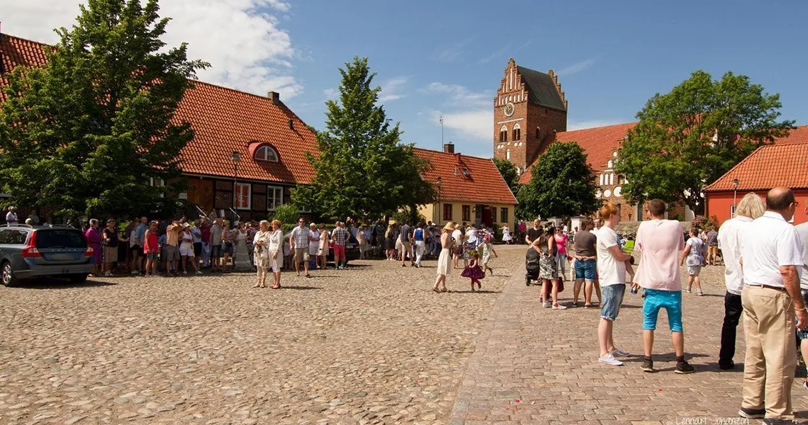 Folk samles på et brostensbelagt torv i Åhus med kirketårnet i baggrunden på en solrig dag