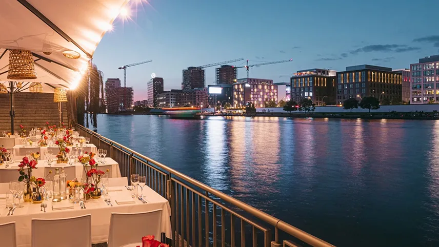Bild från restaurangens vackra terrass medan solen går ner över floden Spree. Borden står dukade med vita dukar, blommor och vinglas.