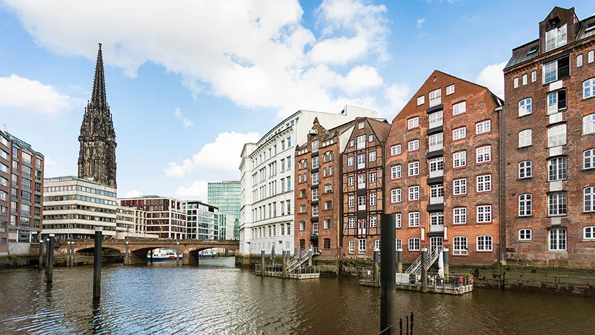Bild från kanalen vid Speicherstadt i Hamburg, med vatten i förgrunden, byggnader, en bro och kyrka i bakgrunden.