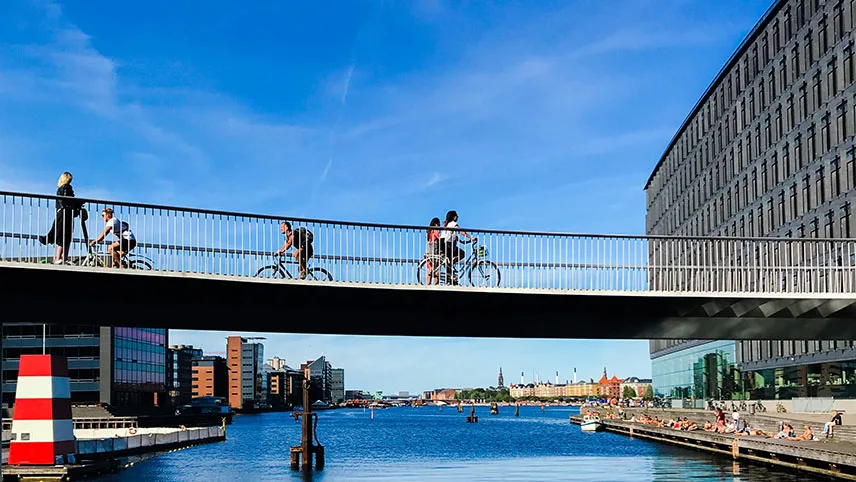 Cyklister på väg över cykelbroen Cykelslangen i centrala Köpenhamn. I bakgrunden syns byggnader och en klarblå sommarhimmel. I förgrunden syns blått vatten och en brygga med badgäster.