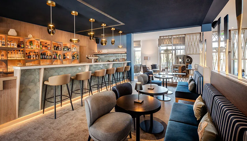 Hotellets bar med en lång bardisk upplyst av lampor, väggfasta soffor och bord.
