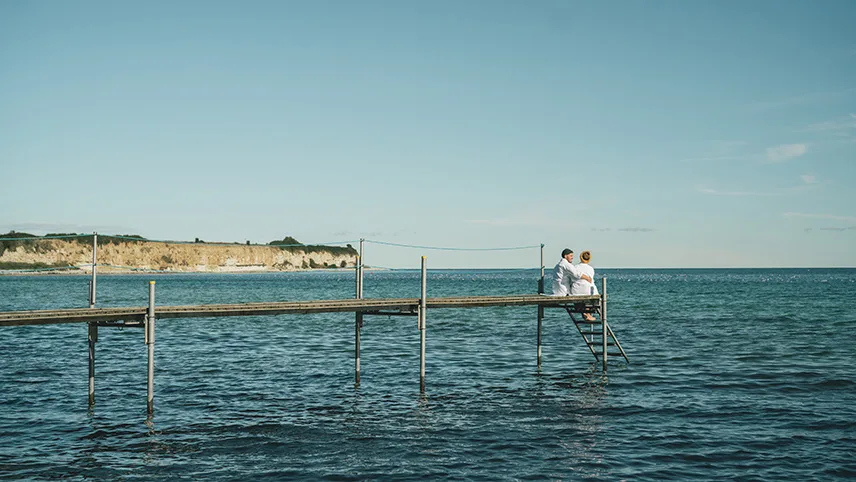 Två personer sitter på en brygga ute på vattnet. De är klädda i vita badrockar och håller om varandra och i bakgrunden syns Stevns Klint.