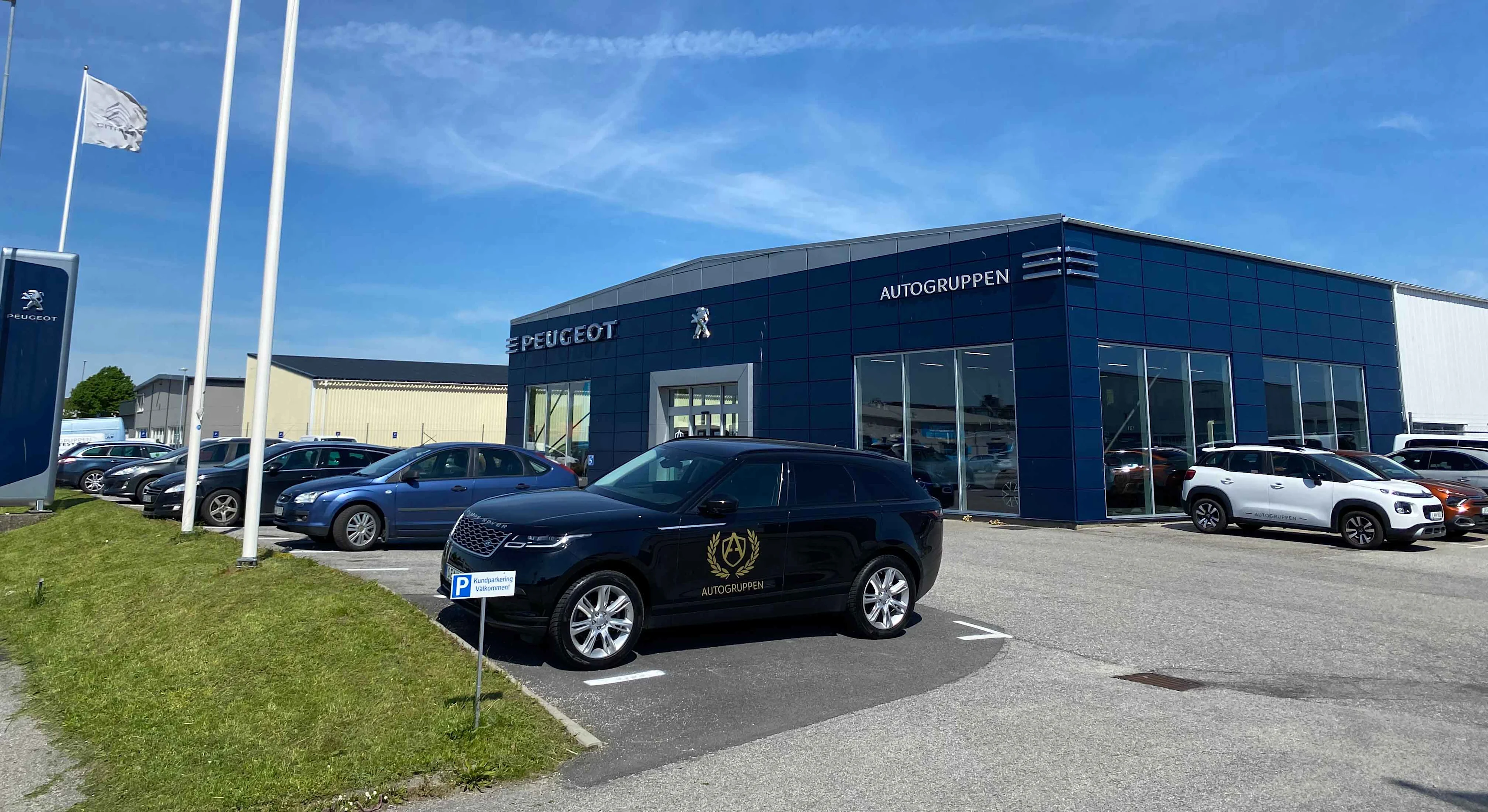 Biler er parkeret foran et bilfirma med navnet "PEUGEOT AUTOGRUPPEN" under en blå himmel med flag i nærheden.