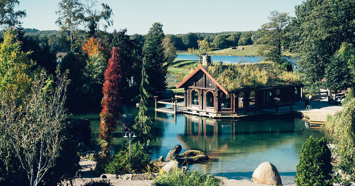Hotelværelser på Ästad Vingård i form af en lille hytte ved vandet med træer ved siden af.
