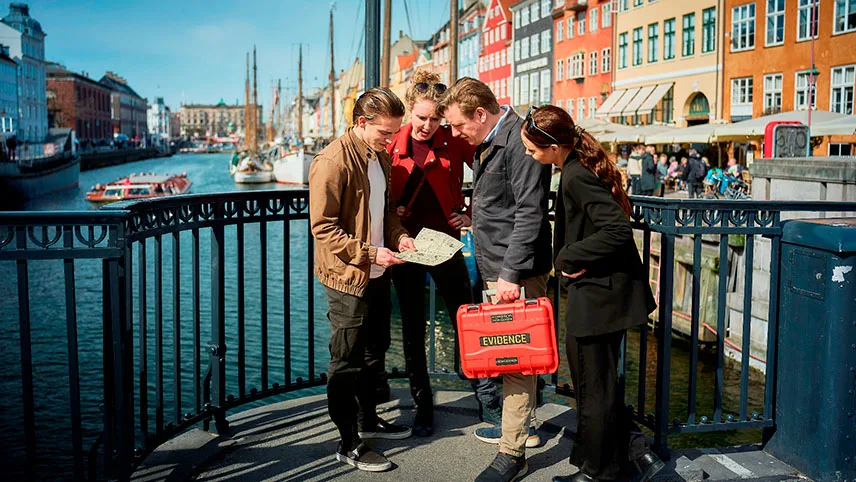 En grupp står på en av gångbroarna i Nyhavn och läser instruktioner. En av besökarna håller i spelväskan och i bakgrunden syns Nyhavns vatten, båtar och färgglada hus.