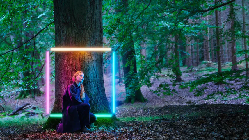 Ett fotografi från utställningen. På bilden syns en grön lövskog. En kvinna sitter på huk framför en trädstam och runt henne lyser en kvadrat bestående av neonljus i fyra olika färger.