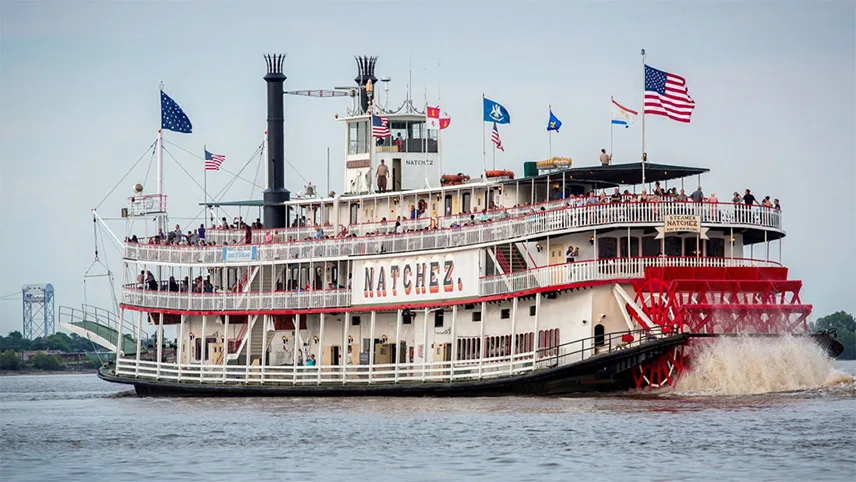 En sightseeing-båt i tre våningar seglar utanför New Orleans i Louisiana. På däck syns resenärer som står och tittar ut och på taket vajar olika flaggor.