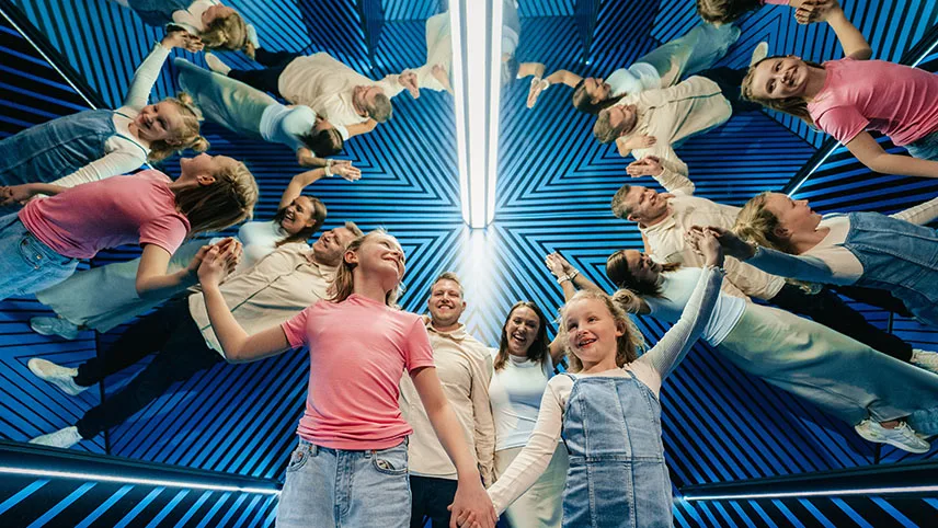En familj befinner sig i ett av illusionsrummen med blå bakgrunden. Perspektivet får det att se ut som att de är väldigt många istället för bara fyra personer.