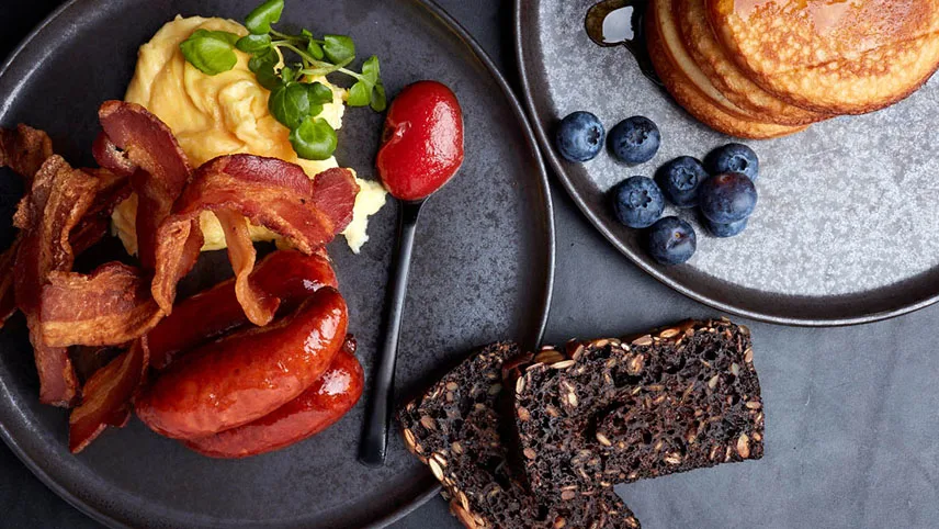 Bild från hotellets frukost. En tallrik med ägg, bacon, korv och rågbröd samt en tallrik med amerikanska pannkakor och färska blåbär.