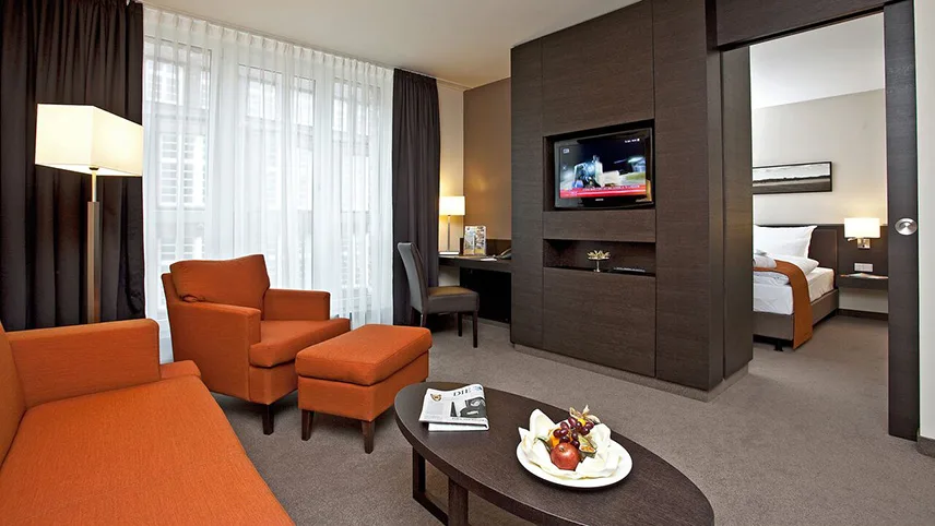 En av sviterna på ATLANTIC Hotel Lübeck. I förgrunden syns en röd-orange sittgrupp med ett soffbord samt tv och skrivbord. I bakgrunden ser man dörren in till sovrummet med en dubbelsäng och en tänd lampa.