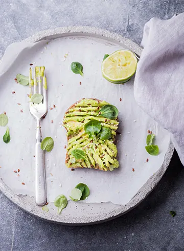 Avokadotoast serverad på en tallrik med limehalva och gröna blad, dekorerad med en gaffel vid sidan, på ett rustikt underlag.