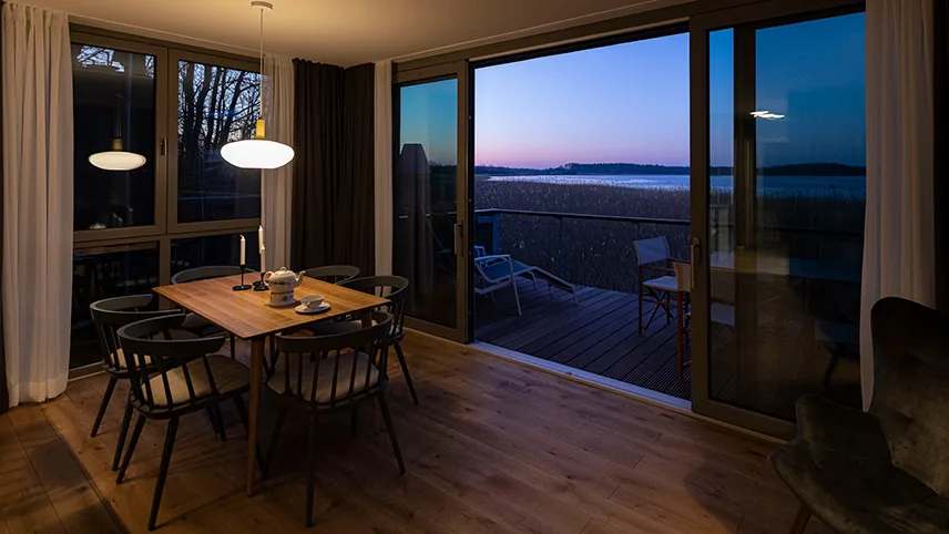 Matplats i ett semesterhus med ett bord och fyra stolar, en tänd lampa samt stora fönster ut mot skymningen.