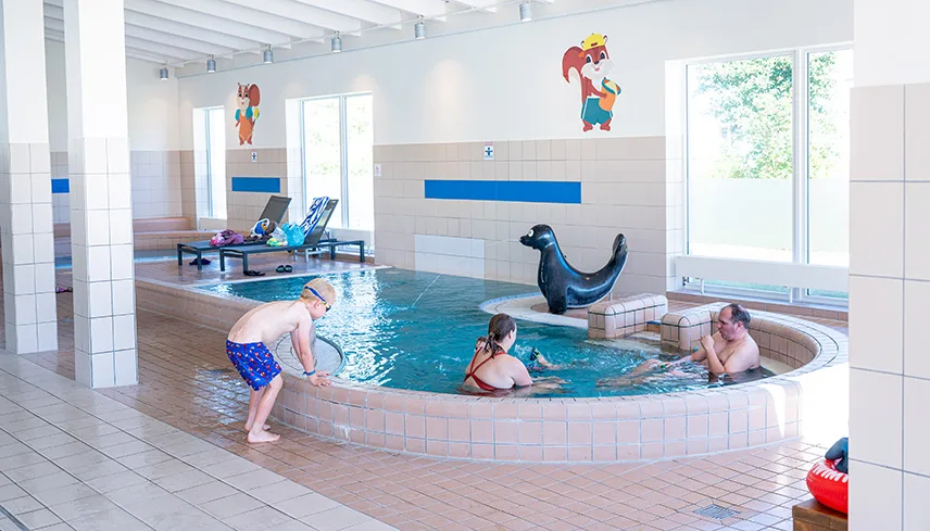 Bild från barnpoolen inomhus. En familj plaskar och leker och man ser solstolar i bakgrunden.