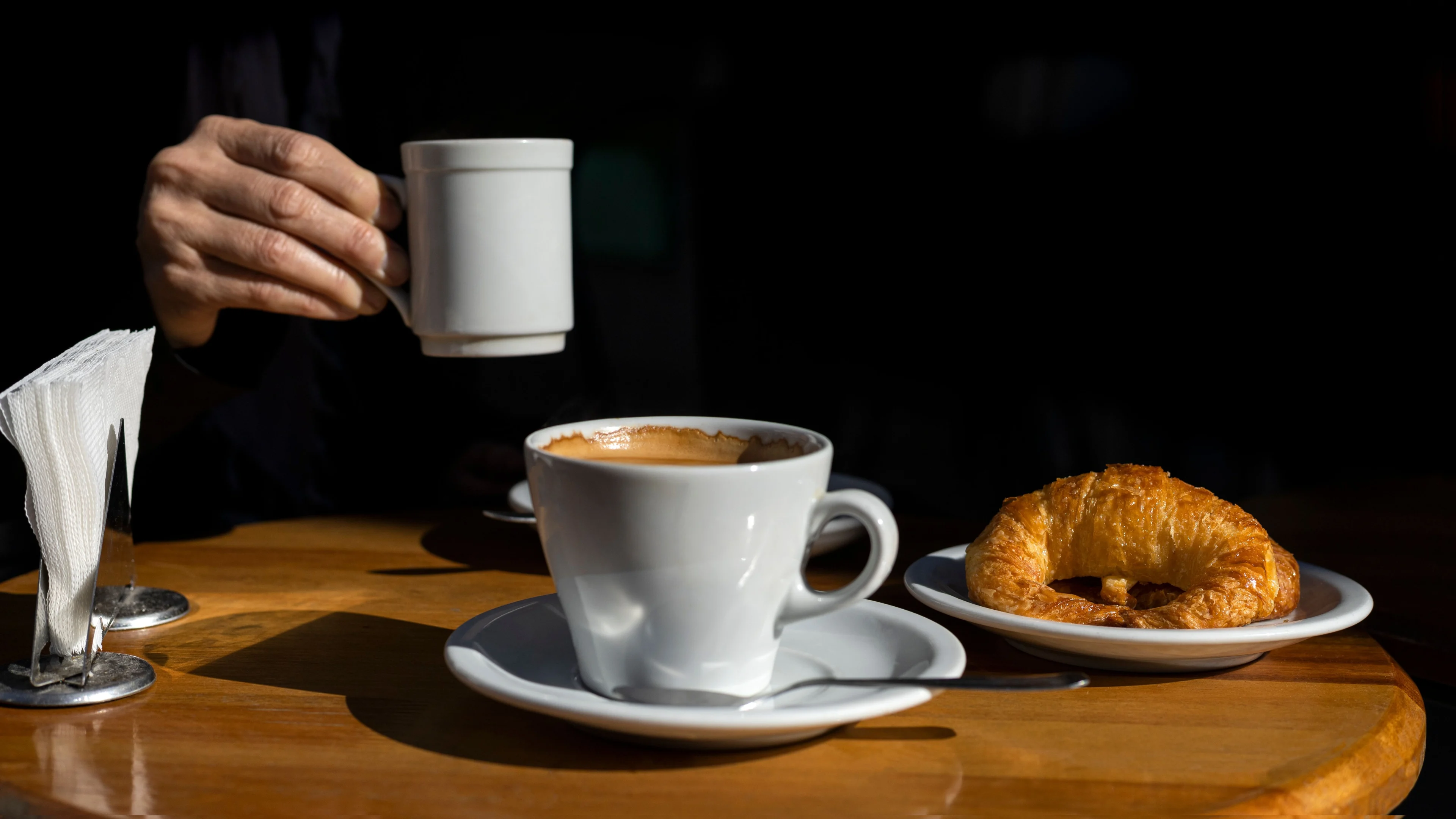 Människa som håller en kopp kaffe i handen och har en croissant framför sig.