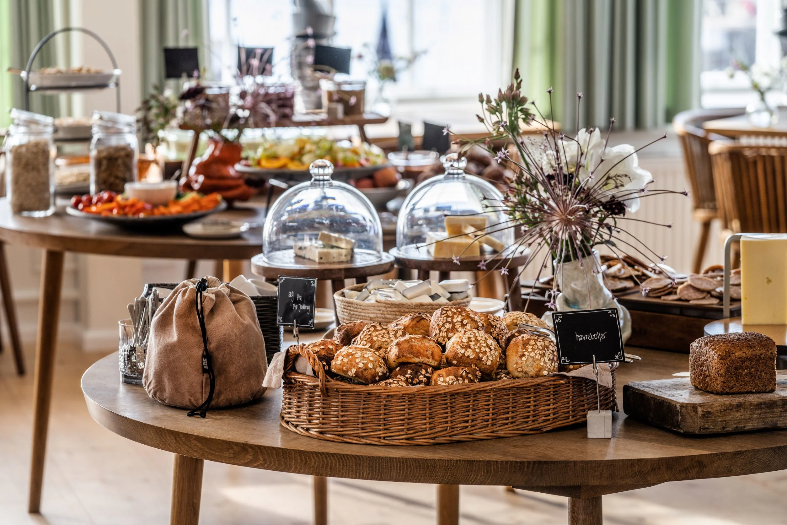 En buffet fyldt med brød, ost og andet bagværk er stillet op på et rundt træbord i et lyst og rummeligt lokale.