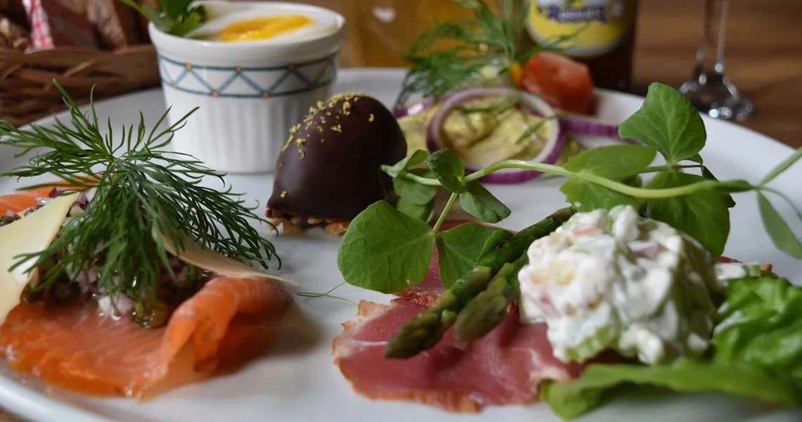 En tallrik med ett urval av aptitretare inklusive lax, sparris, en chokladöverdragen godbit, en kopp äggkräm på ett träbord med en dryck i bakgrunden.