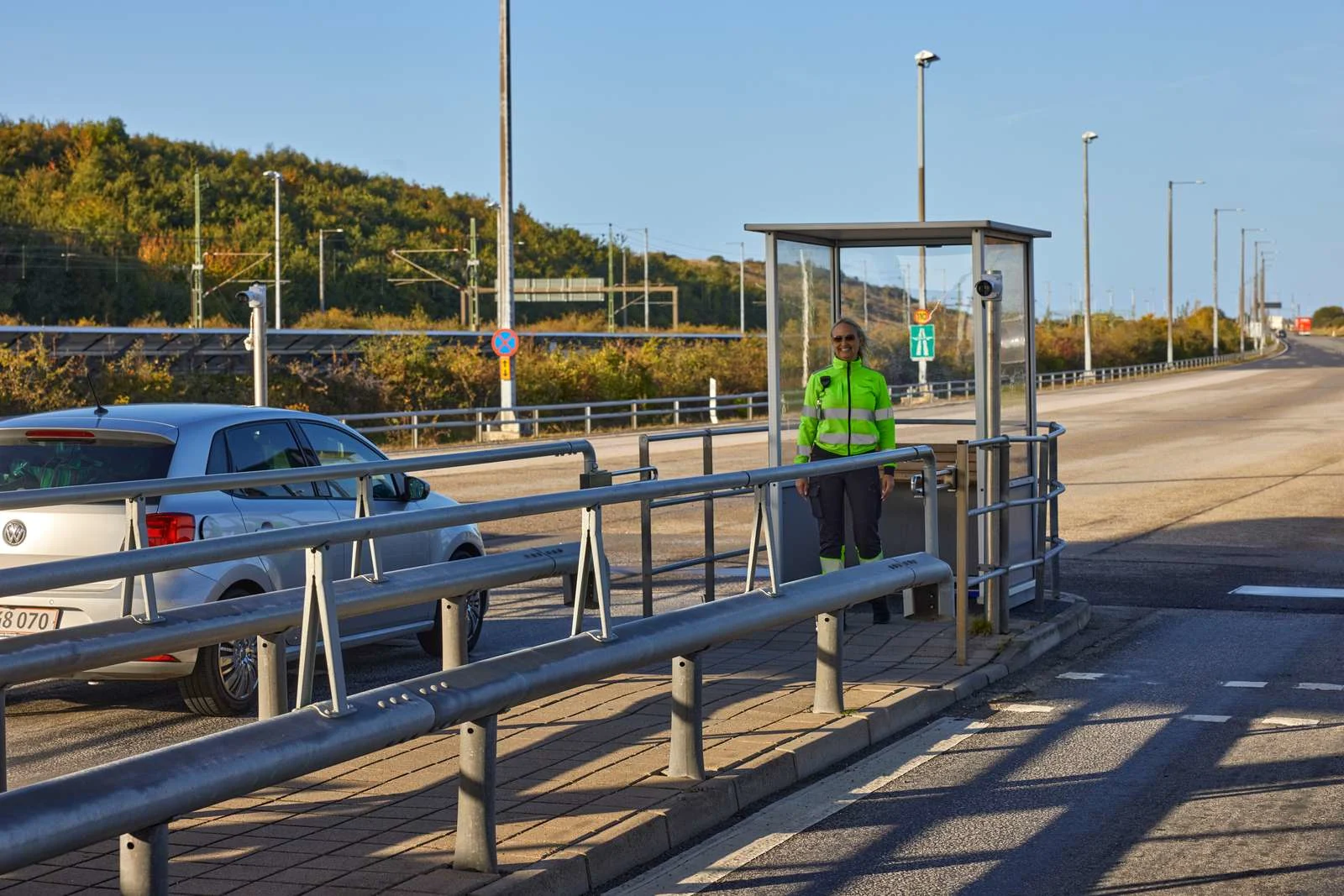 A smiling customs station worker at the Øresund Bridge stands at the payment station while a car waits