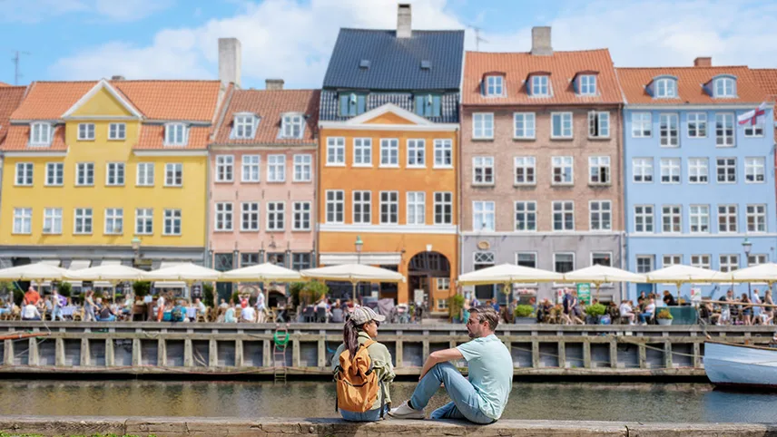 Nyhavn i Köpenhamn med färgglada stenhus och massor av uteserveringar. På kajkanten i förgrunden sitter två människor och pratar.