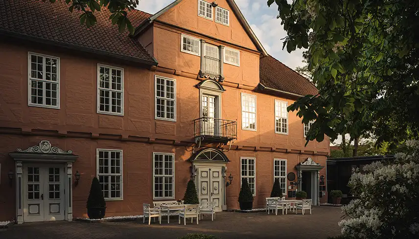 Hotellets fasad sedd snett framifrån. Byggnaden är i barockstil med vita detaljer, framför huset står vita trädgårdsmöbler och grönskande träd ramar in fasaden.