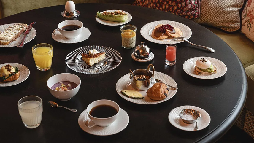 Ett urval av rätter från hotellets frukostbuffé står uppdukade på ett runt bord. Man ser bland annat amerikanska pannkakor med Nutella, Egg Bénedicte, surdegsbröd med smör, en kopp kaffe och ett glas apelsinjuice.